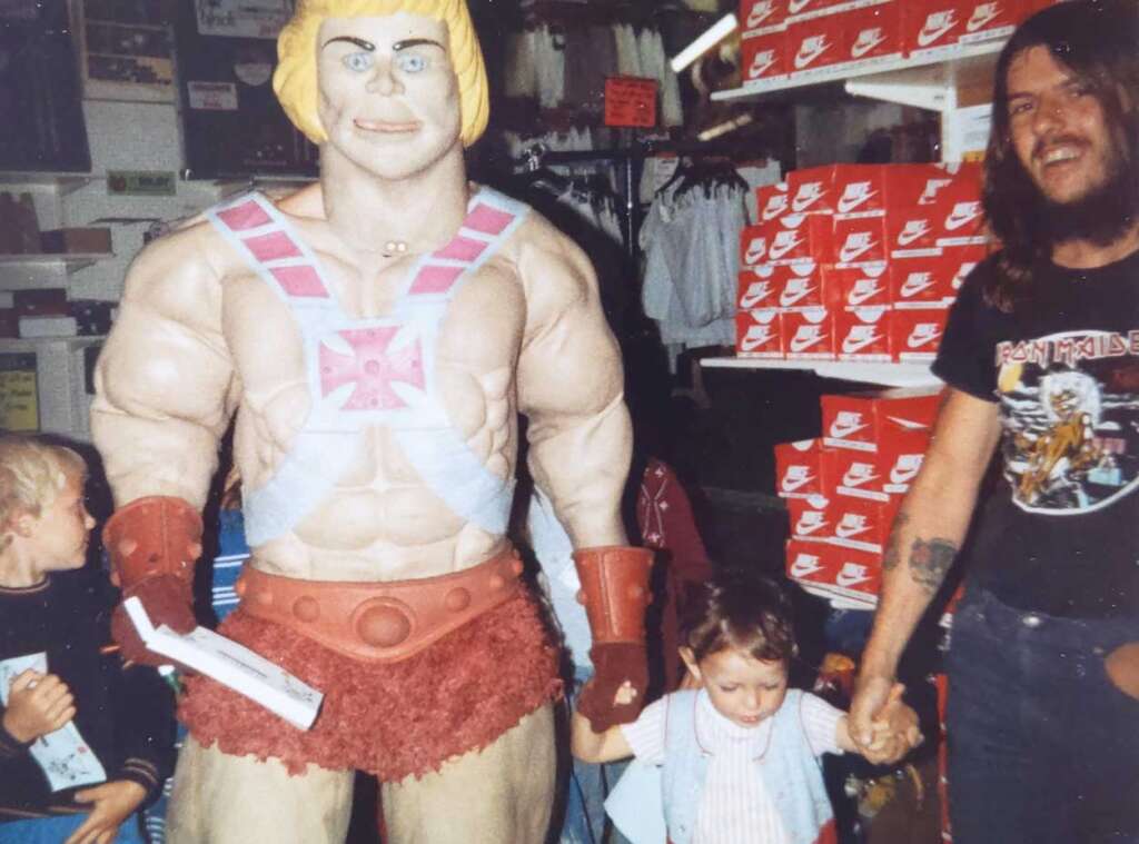 A man holding a child's hand poses next to a large He-Man statue in a store. The child is holding a backpack. In the background are shelves stacked with red Nike shoe boxes. Another child is partially visible, looking at the statue.