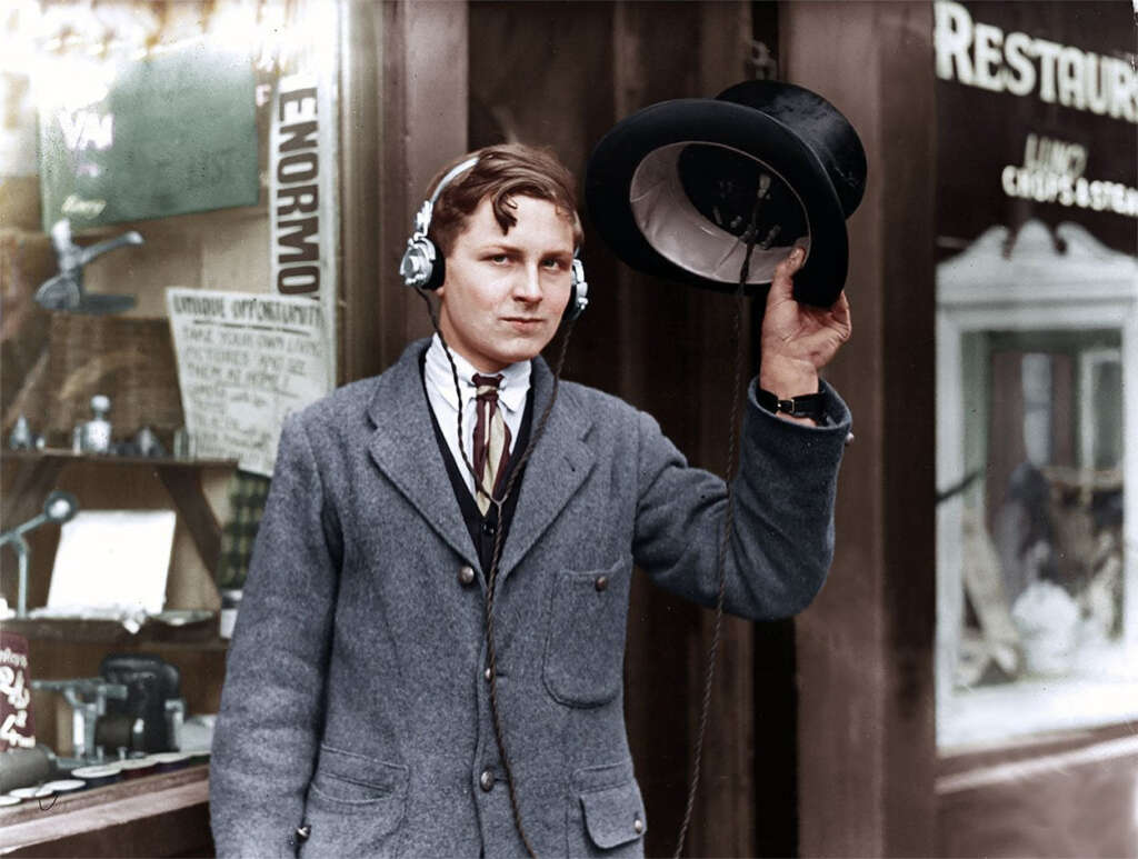 A young man stands outside a shop wearing headphones connected to a radio inside his hat. He is dressed in a vintage suit and tie, gazing slightly to the side. The shop window displays various items and signs.