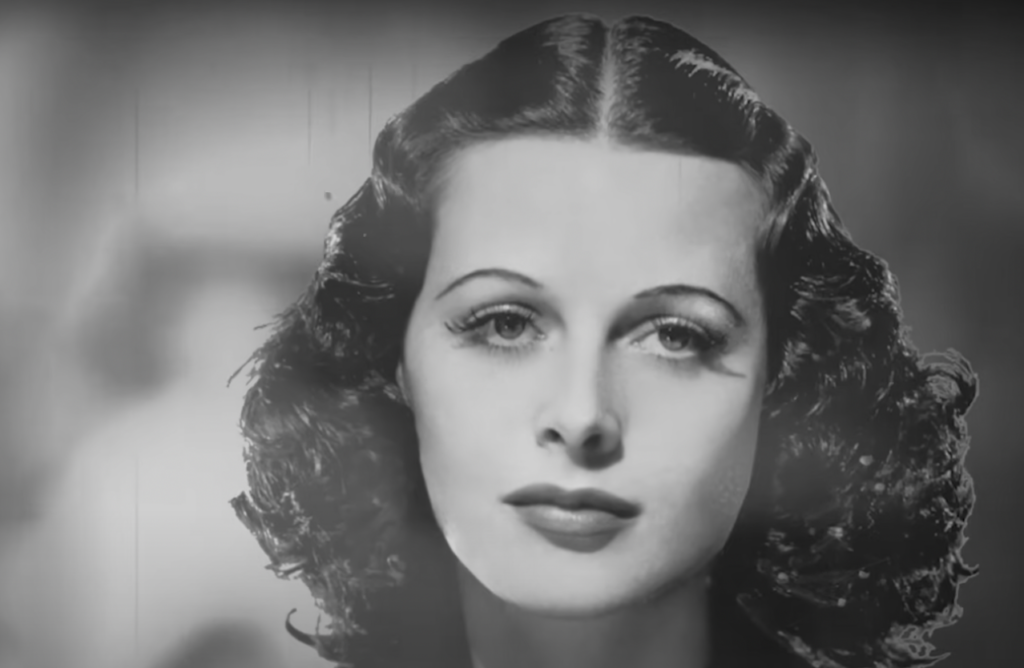 Black and white portrait of a woman with wavy, dark hair parted in the middle. She has a calm expression, arched eyebrows, and is facing the camera directly. The background is blurred, focusing attention on her face.