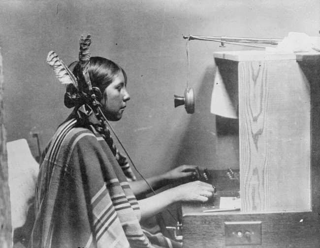 Black and white photo of a young Native American woman with braided hair and feathered headdress using a telephone switchboard. She wears a patterned shawl and sits in profile, operating the equipment.