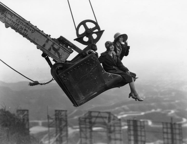 Two women sit on the arm of a large construction crane, high above the hills, wearing dresses and cloche hats, with one woman waving. The scene is vintage, in black and white, with distant hills in the background.