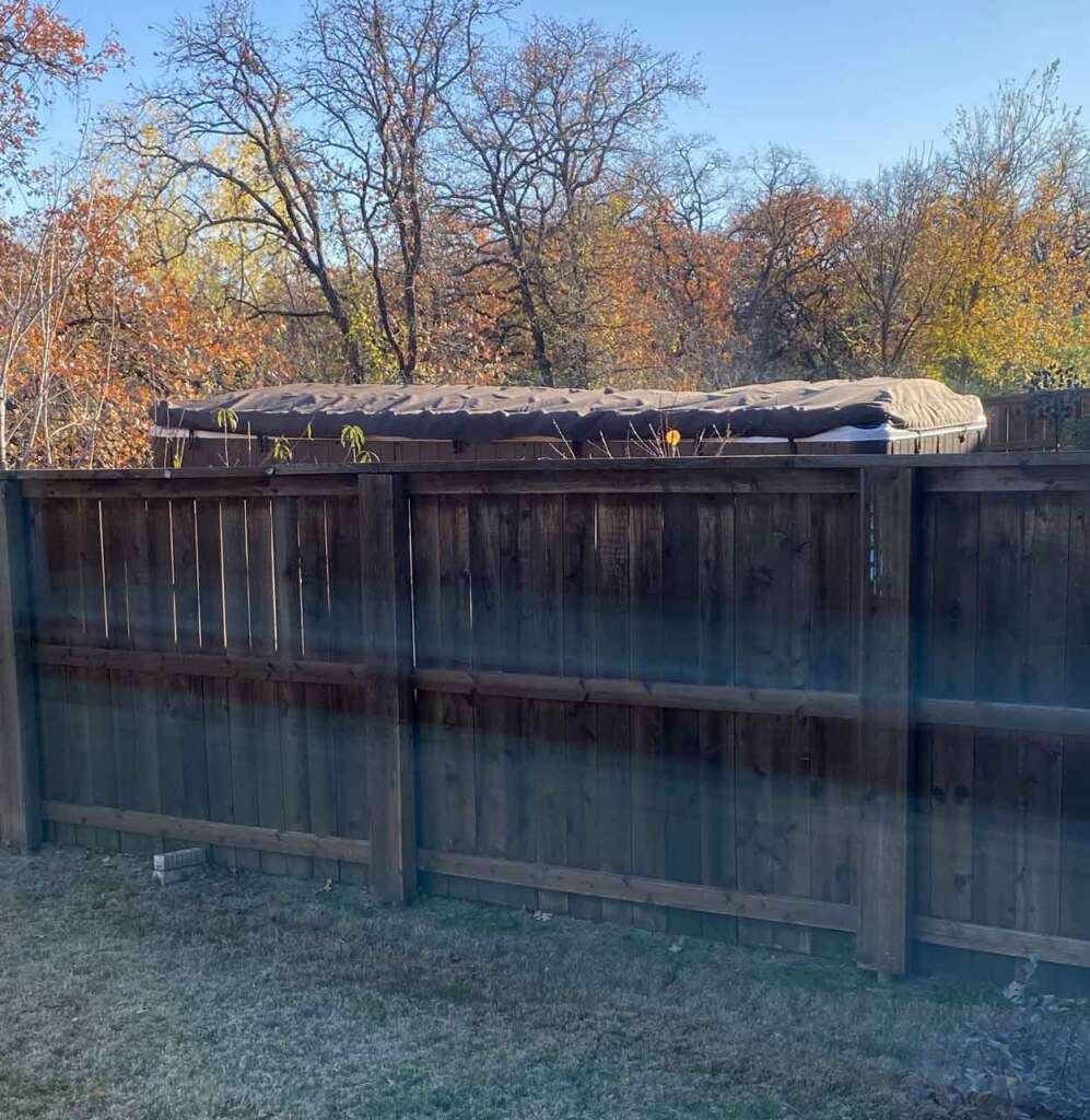 A hot tub with a cover is placed near a wooden fence. The background shows trees with autumn leaves under a clear blue sky. The early morning sun casts soft light and shadows.