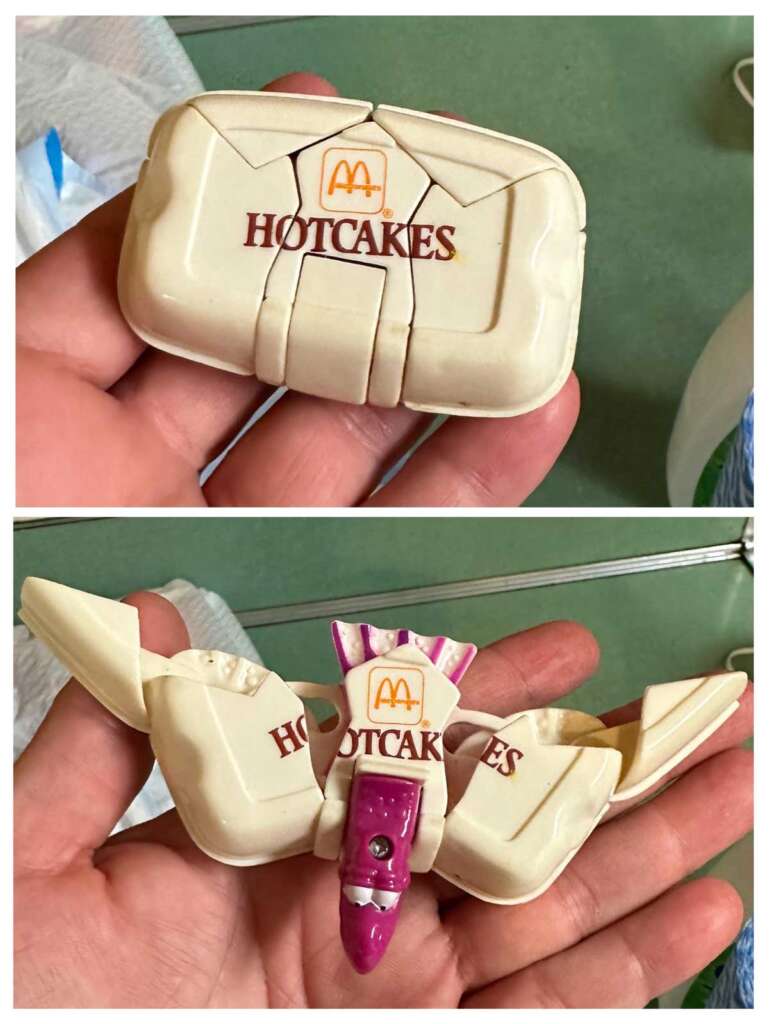 A hand holding a vintage toy shaped like a closed fast-food hotcakes container with an 'M' logo on the top. The toy transforms into a pink and white bird-like creature, with the wings displaying the word "HOTCAKES.