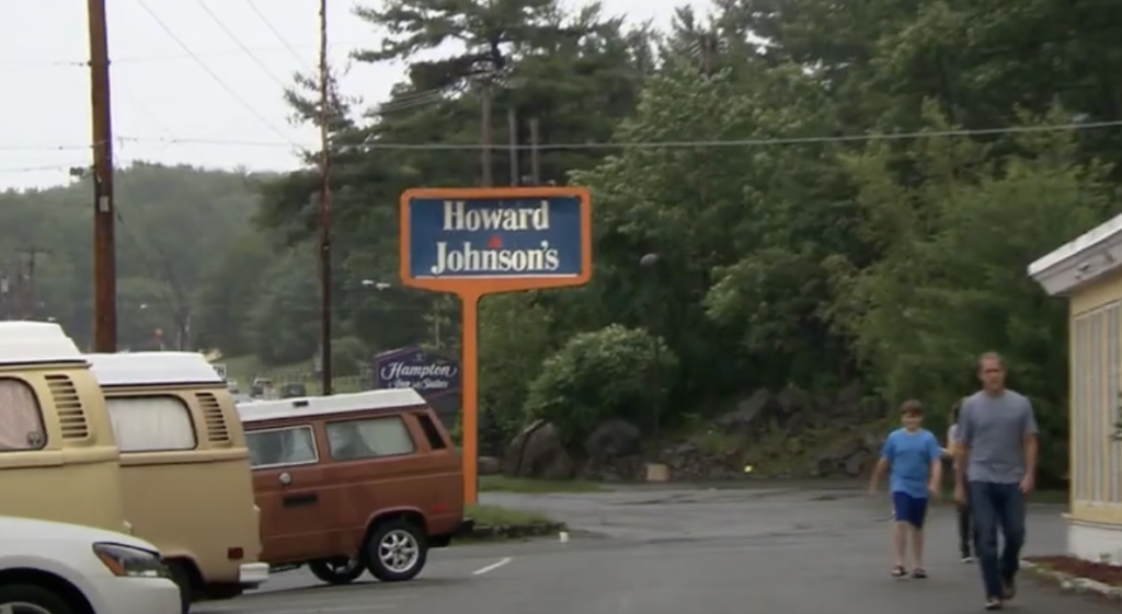 A parking lot with several vans is adjacent to a Howard Johnson's sign. Two people are walking on the right side. Trees and a gray sky are in the background.