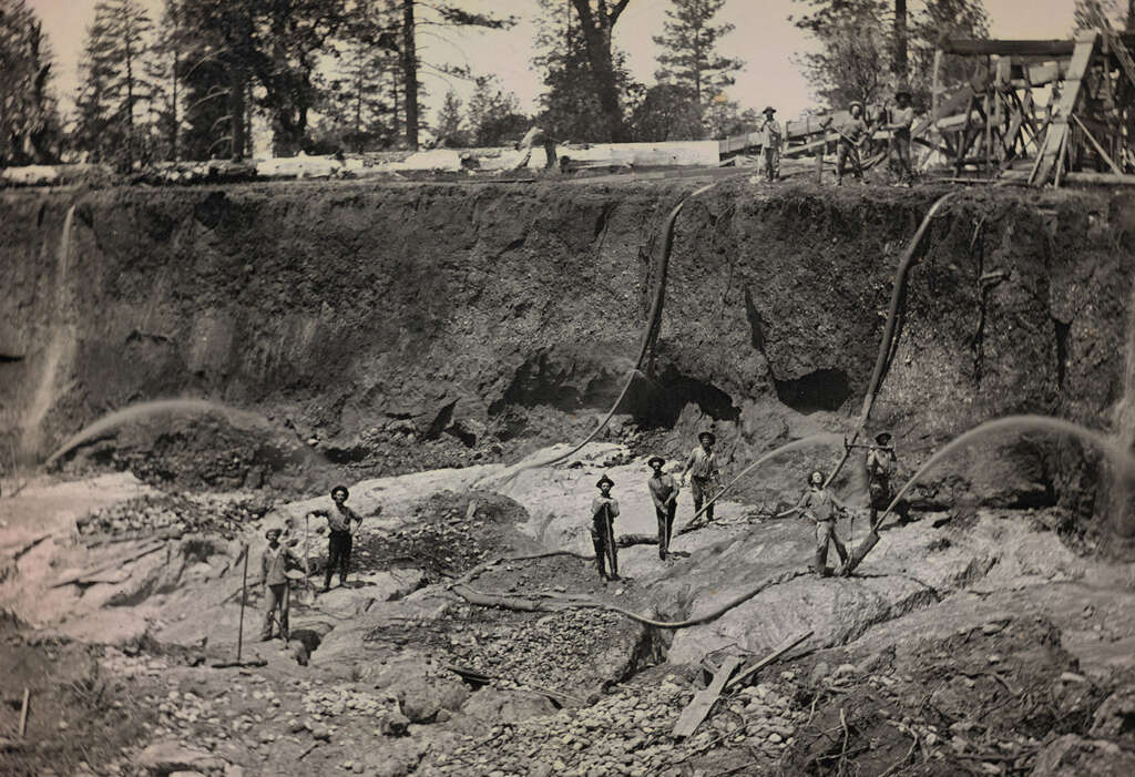 Historic image of several miners using high-pressure water hoses for hydraulic mining in a large pit. The eroded earth and surrounding trees are visible, with wooden structures and workers at the top edge.