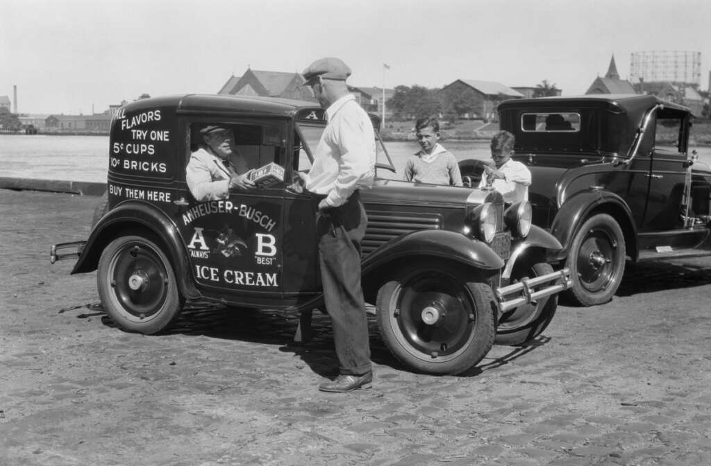 A vintage black-and-white photo depicting a man at an ice cream truck with "Anheuser-Busch Ice Cream" and pricing details on its side. A man in a shirt and cap serves two young boys in the background. Vintage cars are parked nearby.