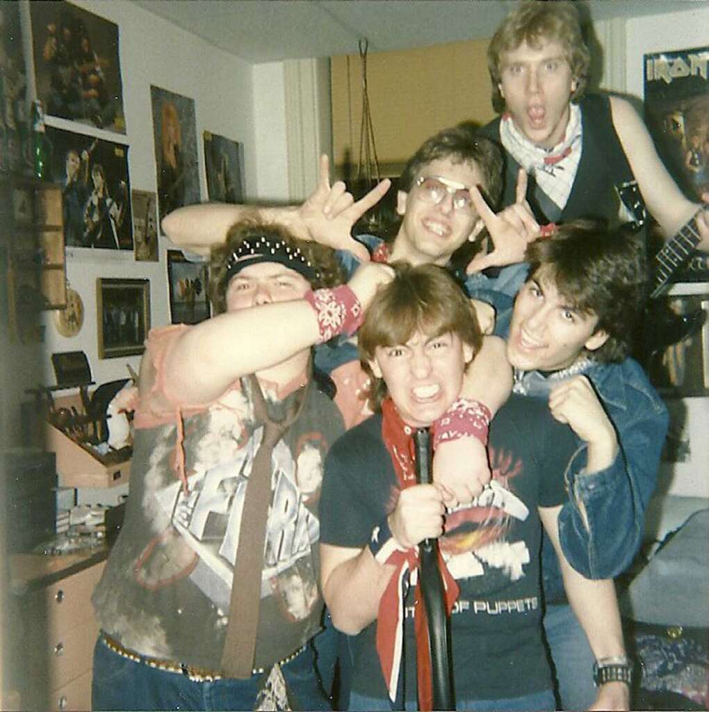 Five people in a room are posing energetically, dressed in casual 1980s-style clothing. One person is holding a microphone stand. Posters and photos are visible on the walls, and the group appears excited and lively.