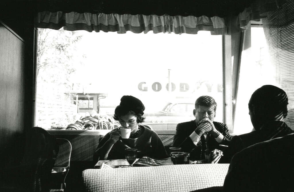 Black and white photo of three people seated in a diner. Two are sipping from cups, facing the window labeled "GOOD Y." Outside, a vintage car is visible. Interior details like curtains and seating are prominent, evoking a mid-20th-century atmosphere.