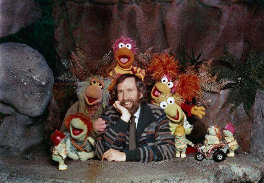 A man with a beard is surrounded by five colorful puppet characters, each with distinct features and expressions. They are in a natural-looking setting with rocks and plants, creating a playful atmosphere.