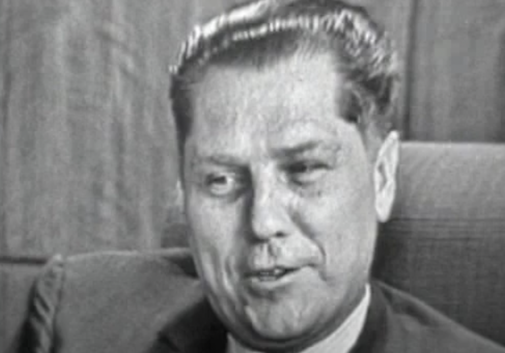 Black and white photo of a man wearing a suit, sitting and speaking. His hair is neatly styled, and he appears to be in a formal setting. The background is blurred, suggesting an indoor environment.