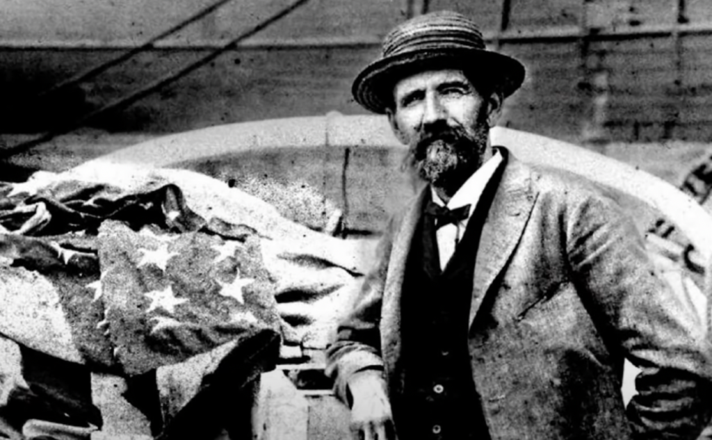 A vintage black-and-white photograph of a bearded man in a suit and bowler hat, standing next to stacked fabric with stars. He leans casually on the fabric, with a ship's deck or similar structure in the background.