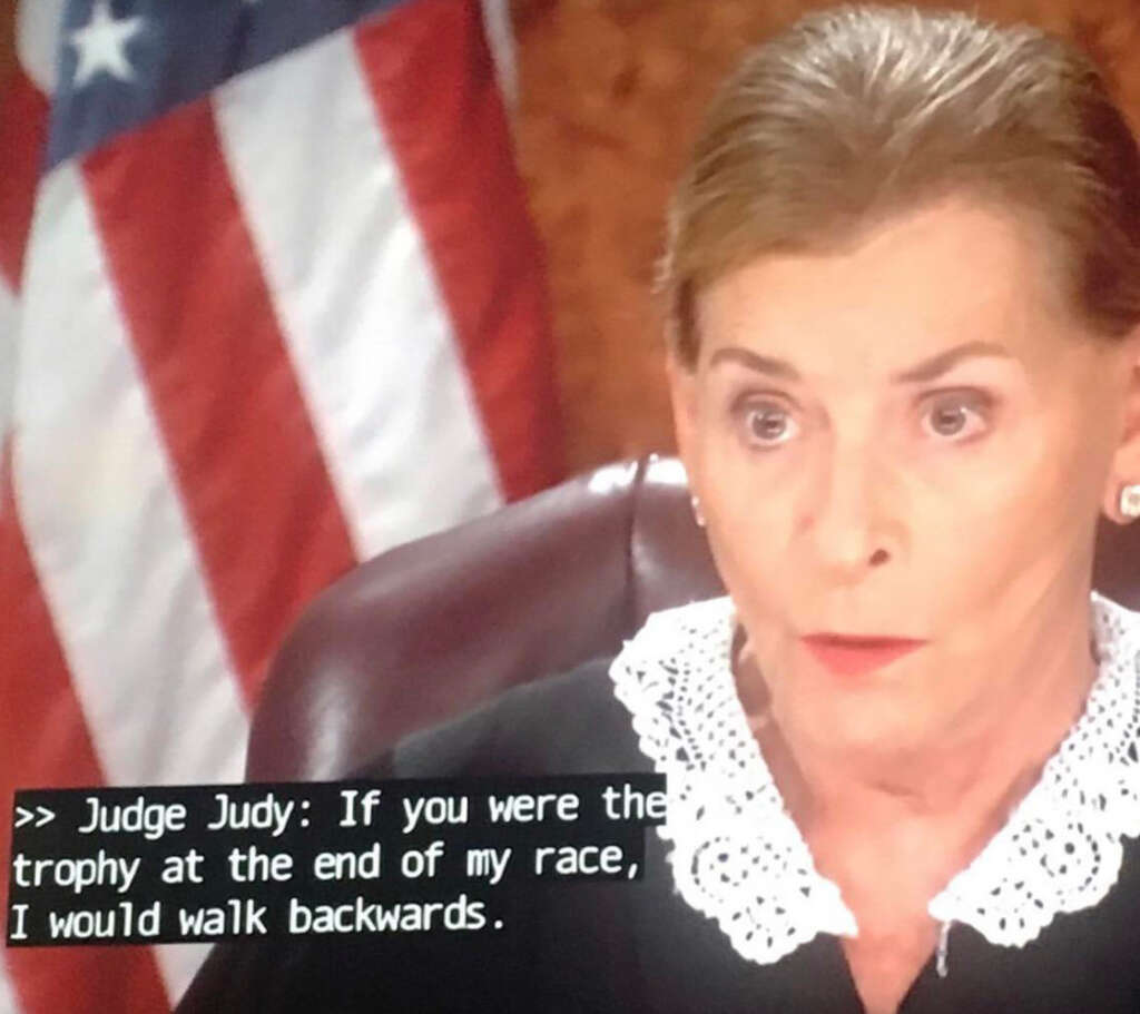 A woman with a stern expression is sitting in a courtroom. She wears a black robe with a white lace collar. An American flag is partially visible in the background. Subtitles read: "If you were the trophy at the end of my race, I would walk backwards.