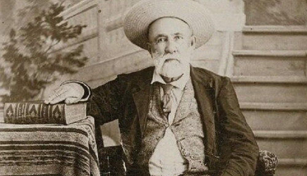 An elderly man with a beard and mustache, wearing a straw hat and suit, sits beside a table with a large book. The setting appears to be indoors with a backdrop of stairs and a potted plant. The image is in sepia tone.