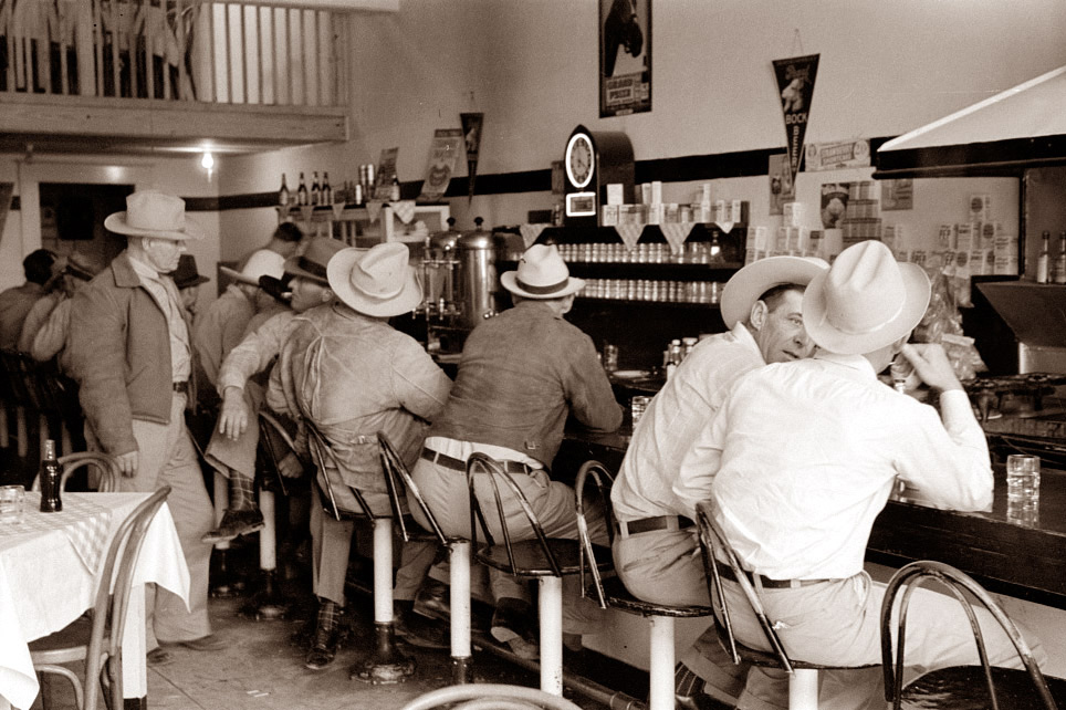 Black-and-white photo of men in cowboy hats sitting at a busy diner counter. They are engaged in conversation, with a jukebox nearby and vintage advertisements on the walls. An upper loft is visible in the background.