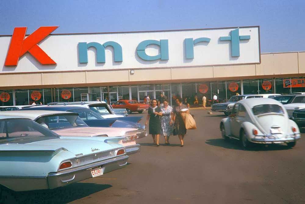 Vintage Kmart Photos- scene of a Kmart parking lot with classic 1960s cars, including a Volkswagen Beetle. Three women, wearing mid-20th century dresses, walk towards the camera carrying shopping bags. The building facade features a large "Kmart" sign.