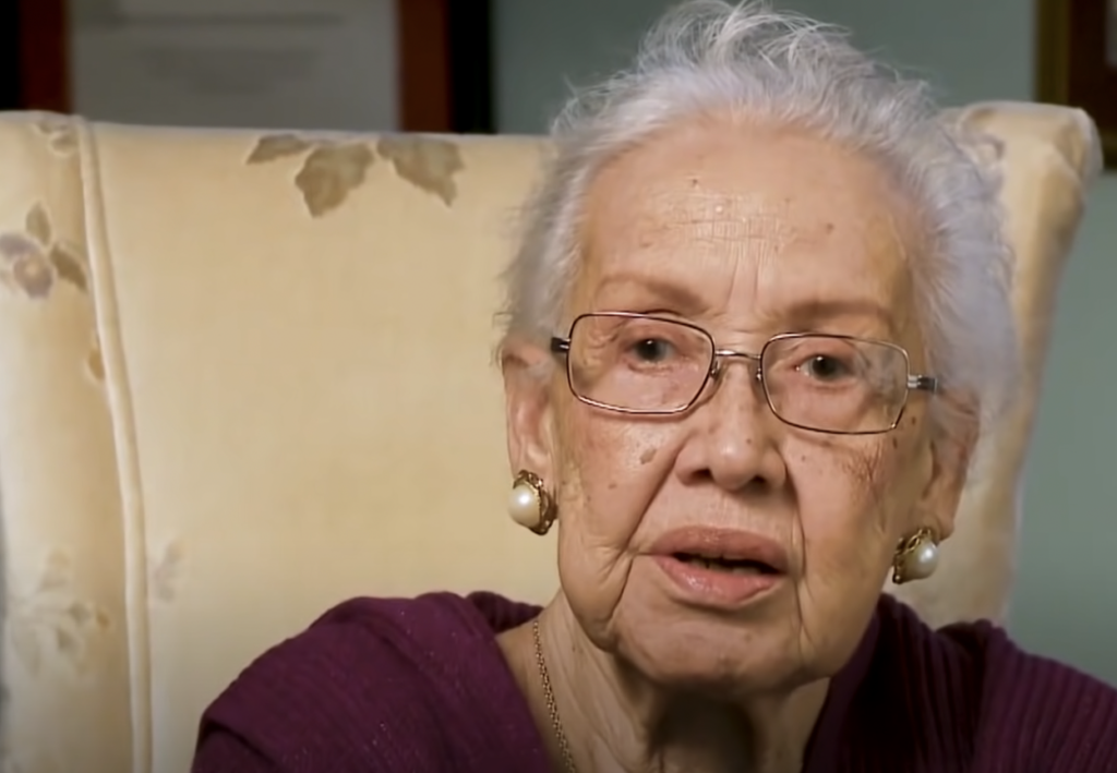 An elderly woman with glasses and white hair sits on a patterned chair. She is wearing a maroon top and gold earrings, looking slightly to the side.