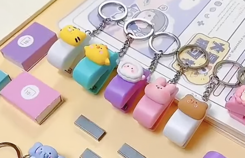 Colorful keychains with cute animal and character designs are displayed on a table. Each keychain features a different character on top of a white clip. Nearby are other small stationery items, including miniature notebooks.