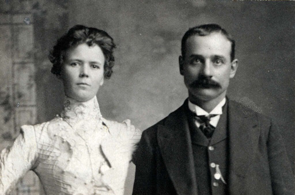 A vintage black and white photograph of a woman and a man. The woman is wearing a high-necked dress with intricate lacework, and the man is in a suit with a bow tie and mustache. They both have neutral expressions.