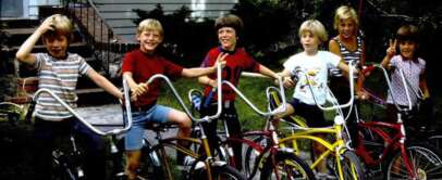 Six children are happily posing with their bikes on a sidewalk in front of a house. The bikes are in different colors, and one child is holding a red flag attached to a bike. They are outdoors, with greenery in the background.