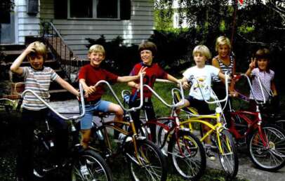 Six children are happily posing with their bikes on a sidewalk in front of a house. The bikes are in different colors, and one child is holding a red flag attached to a bike. They are outdoors, with greenery in the background.