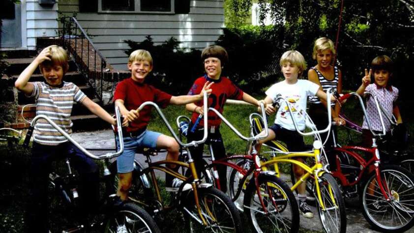 Six children are happily posing with their bikes on a sidewalk in front of a house. The bikes are in different colors, and one child is holding a red flag attached to a bike. They are outdoors, with greenery in the background.