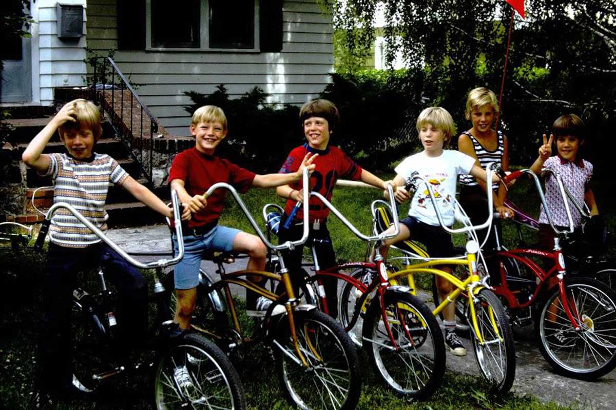 Six children are happily posing with their bikes on a sidewalk in front of a house. The bikes are in different colors, and one child is holding a red flag attached to a bike. They are outdoors, with greenery in the background.