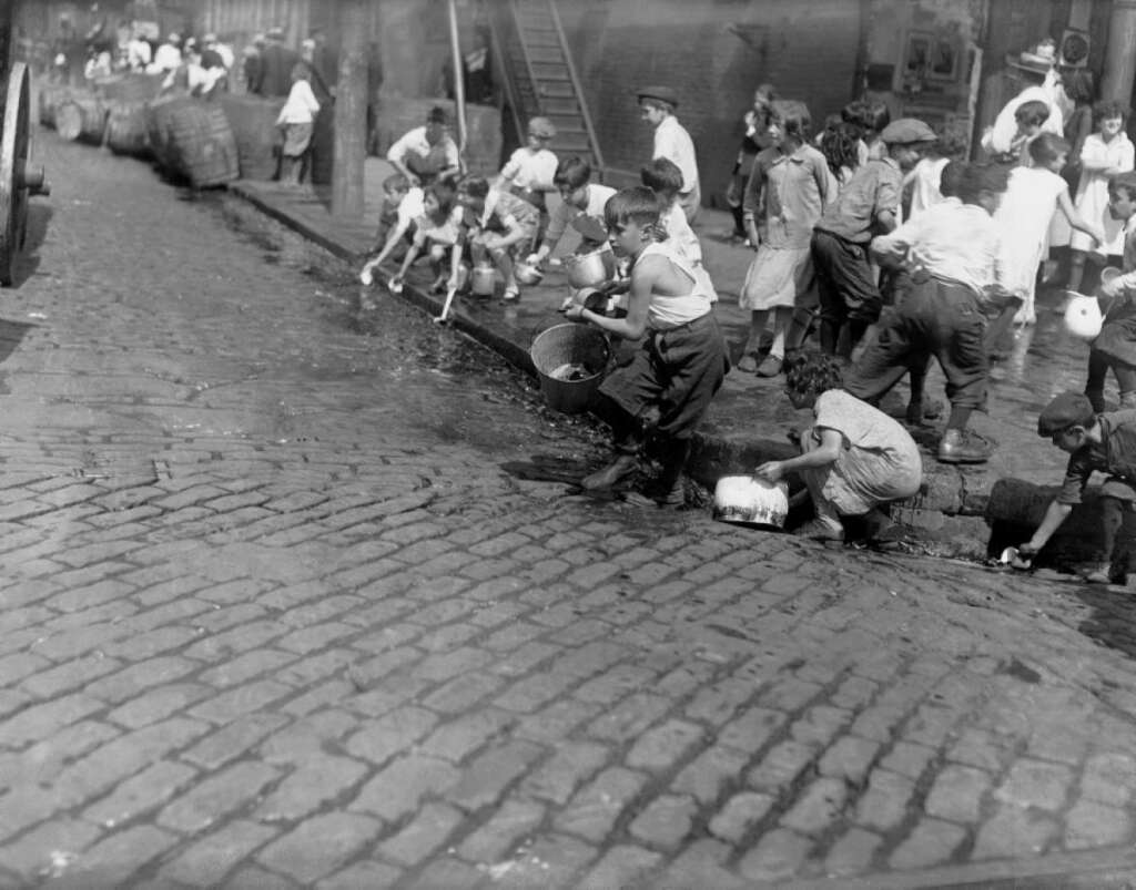 Children playing and gathering water from a gutter on a cobblestone street. Some are seated on the sidewalk with buckets and containers, while others stand or gather in groups. The background shows more children and a few adults near buildings.