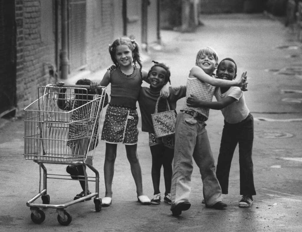 A black-and-white photo of five smiling children standing in an alley with their arms around each other. One child is leaning against a shopping cart. They appear playful and joyful in the urban setting.