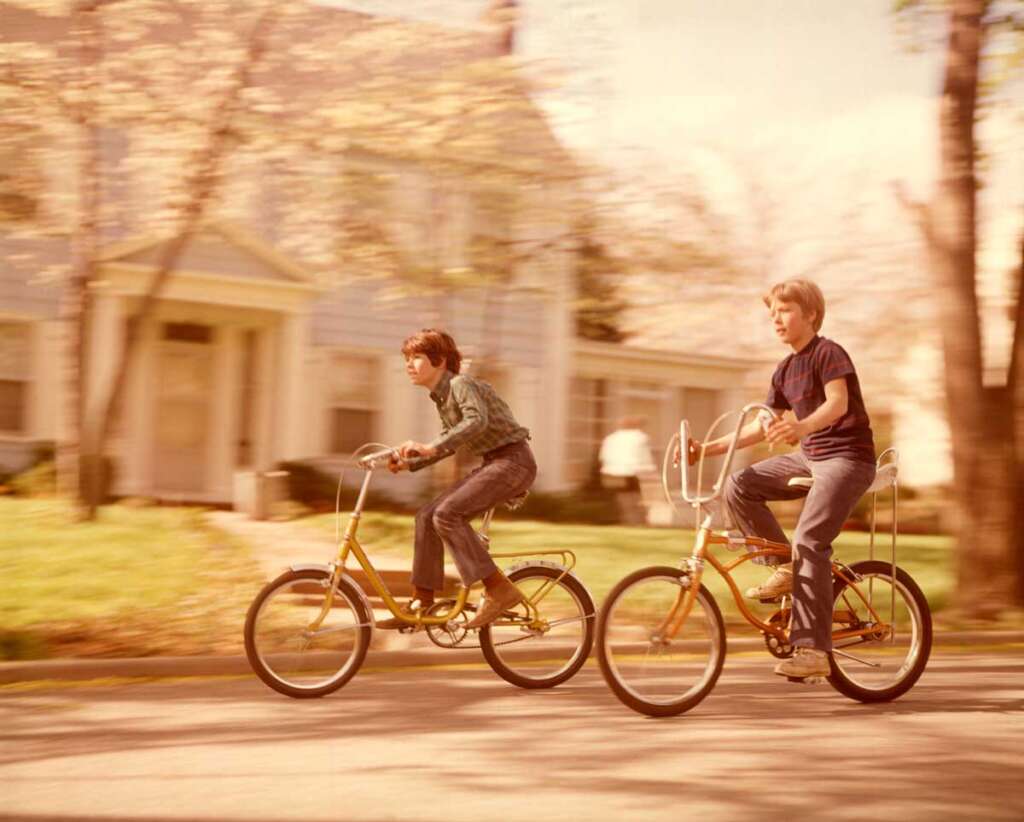 Two boys ride bicycles down a suburban street. The child on the left wears a dark jacket, and the one on the right wears a navy shirt. Both bikes have chrome handlebars and are in motion, with a blurred house and trees in the background.