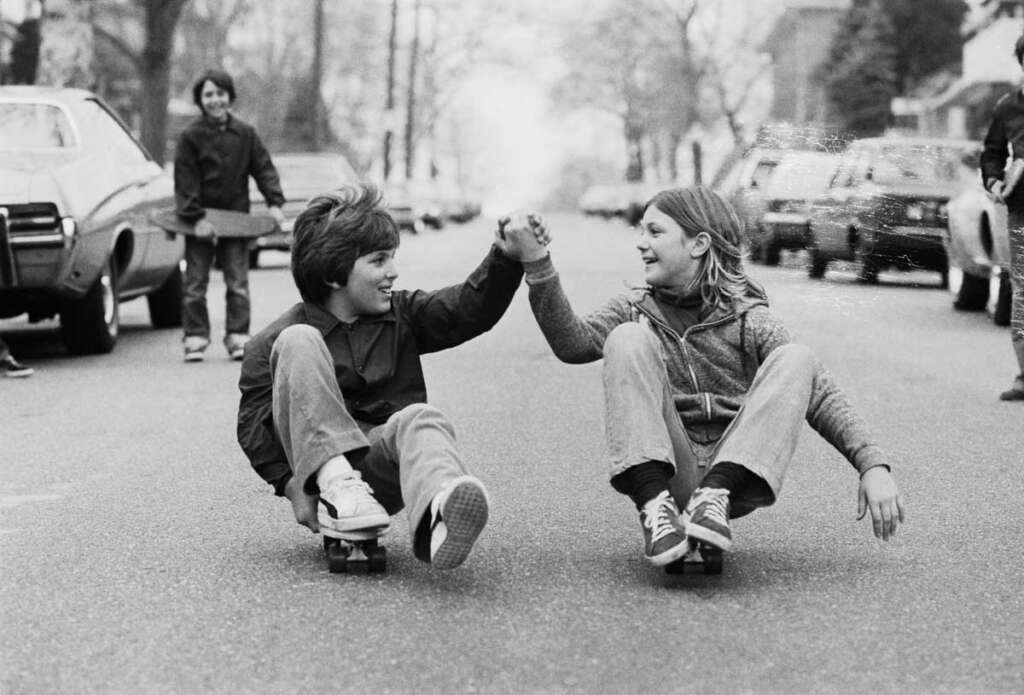 Two kids sitting on skateboards hold hands and smile on a quiet street, while another child stands in the background. Parked cars line the sides of the road. The scene is in black and white, evoking a playful and nostalgic atmosphere.