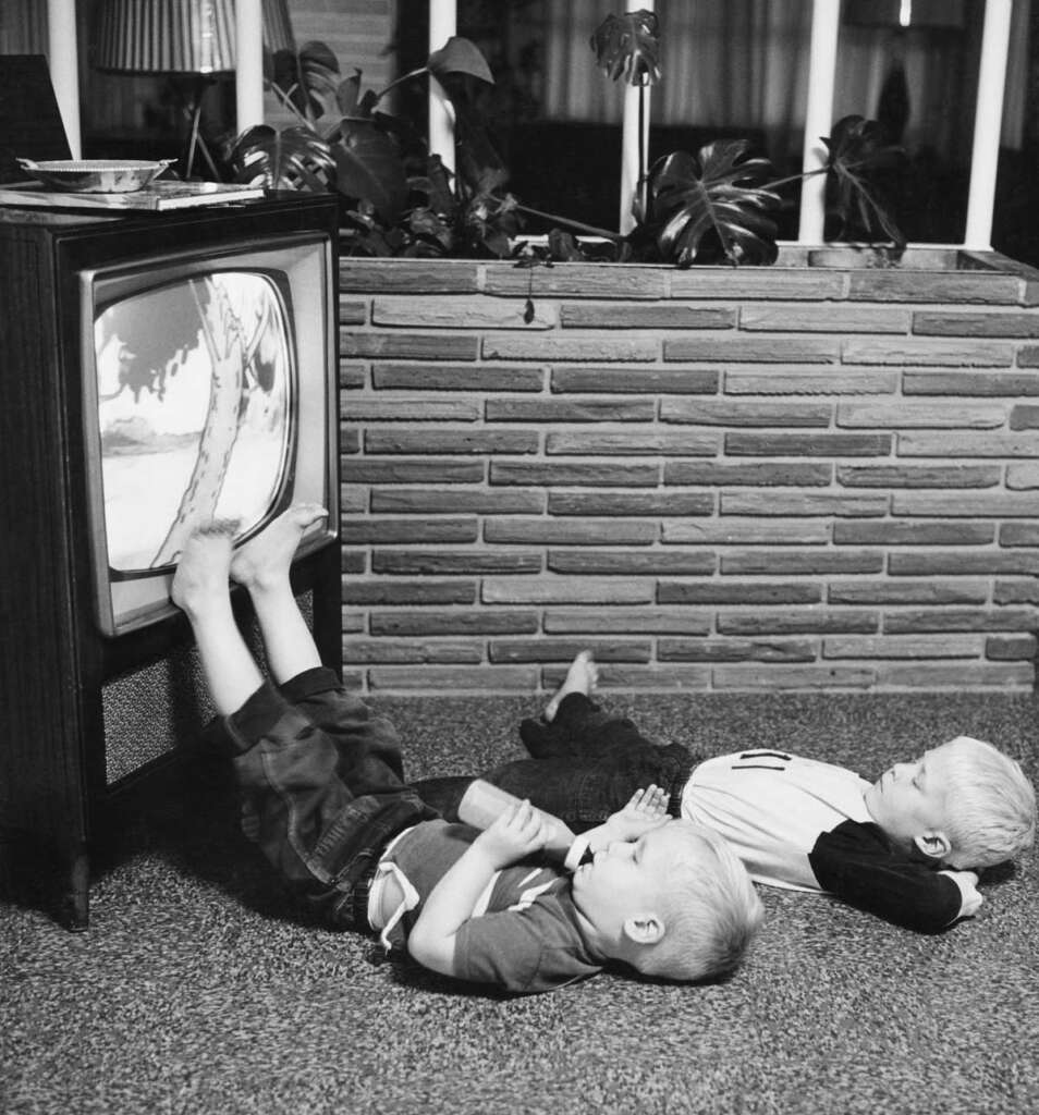 Two children lying on the floor watch a black-and-white television, one with feet resting on the screen. Behind them is a brick wall with potted plants. The scene captures a vintage, cozy indoor setting.