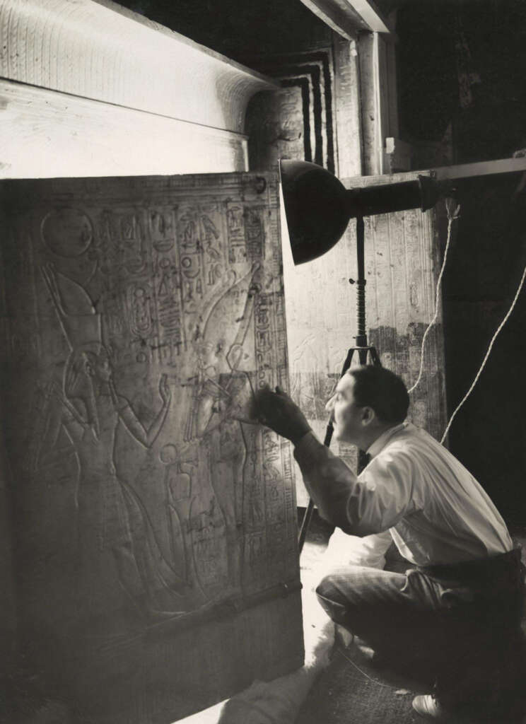 A man carefully examines an ancient Egyptian wall carving illuminated by a spotlight. He crouches, intently studying the intricate hieroglyphic details and figures etched into the stone.