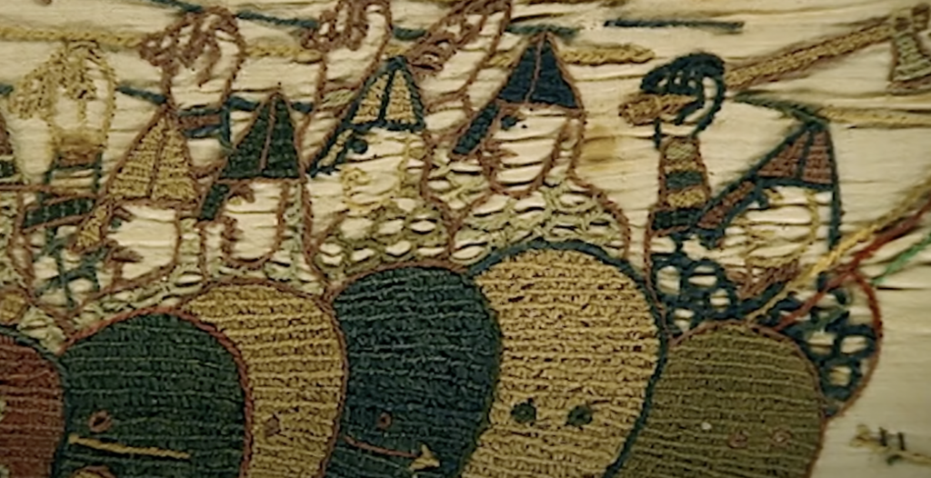 Close-up of a section of the Bayeux Tapestry depicting medieval soldiers. They wear conical helmets with nose guards, carrying shields with various patterns, and their faces have minimal, stylized features.