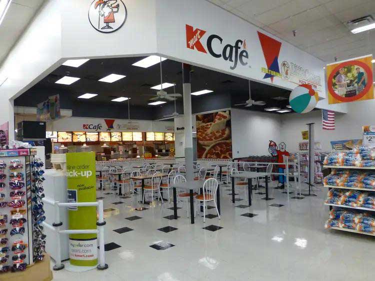 An empty indoor cafe area in a store with white tables and chairs. The walls display images of pizza and the K Cafe logo. Nearby are snack racks and a child ride. The floor is white with black tiles, and the U.S. flag is on the wall.