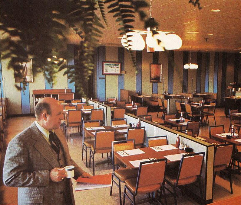 A man in a suit holds a coffee cup, standing in an empty restaurant with neatly arranged tables and chairs. The room features striped walls, pendant lights, and hanging plants, creating a retro ambiance.