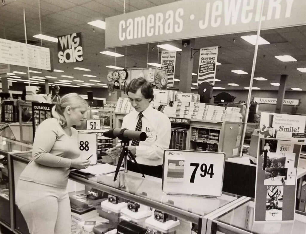 A woman and a man are at a store counter in the cameras and jewelry section. The woman is holding a box. There's a telescope on the counter, price tag visible, surrounded by other electronics. A "WIG SALE" sign is in the background.