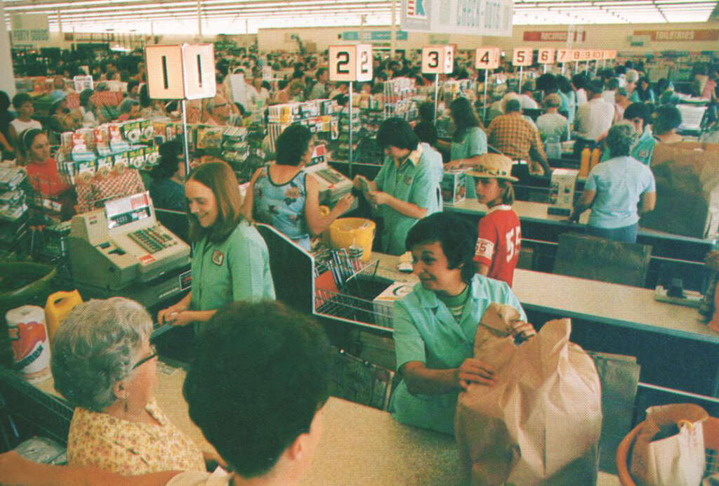 A busy supermarket checkout area with numerous shoppers queuing. Employees in green uniforms assist customers with groceries. A mix of different people and products are visible in a lively atmosphere.