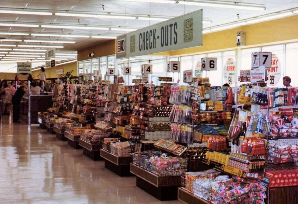 A vintage supermarket checkout area with numbered lanes, stocked with various colorful snacks and candies. Signs hang above the lanes, and shoppers can be seen in the background pushing carts.