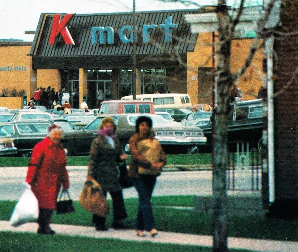 People walk past a Kmart store entrance, with the logo prominently displayed on the facade. Cars fill the parking lot, and several individuals are visible near the entrance. Trees and a walkway are in the foreground.