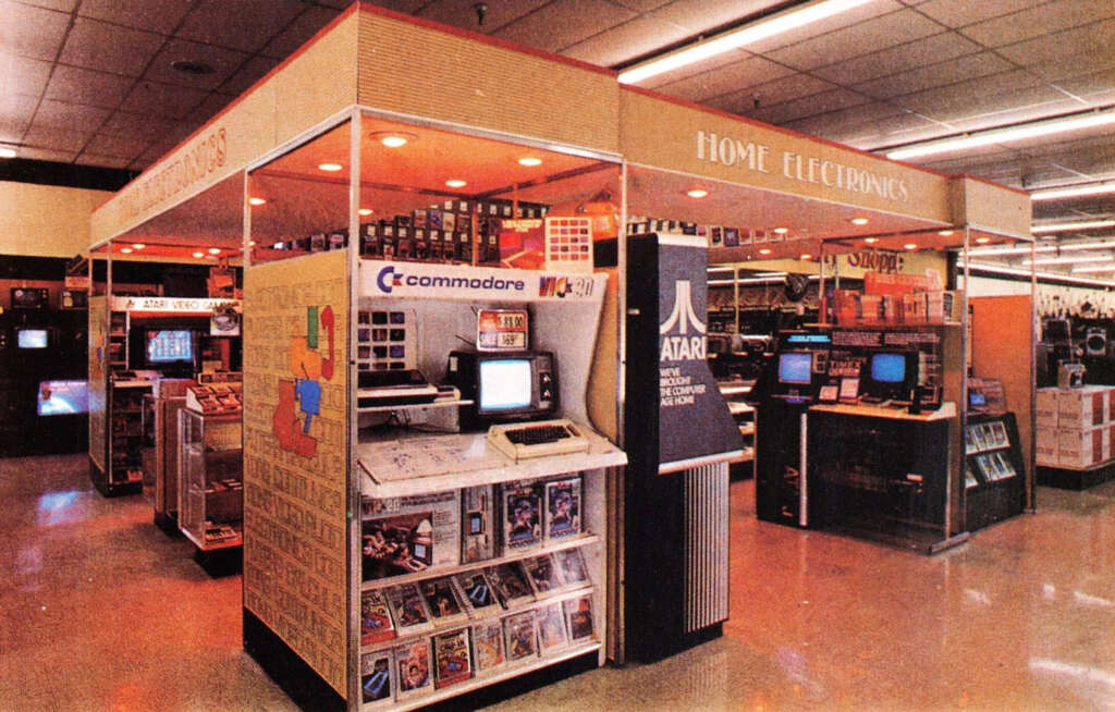 Image of a retro home electronics store featuring vintage computers and arcade machines. Visible brands include Commodore and Atari. Shelves are stocked with software, gaming cartridges, and electronic devices under fluorescent lighting.