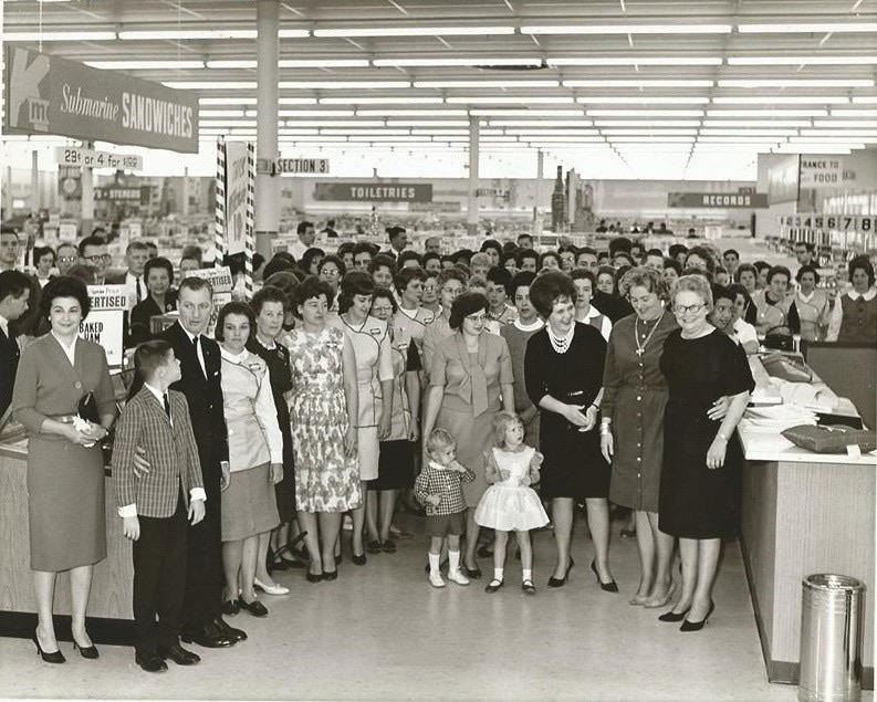 A large group of people, including women, men, and children, are gathered inside a store. The aisle signs indicate sections like toiletries and records. The fashion and decor suggest a mid-20th-century setting.