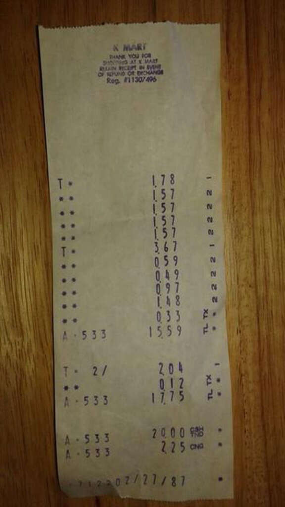 A receipt with faded text lies on a wooden surface. It includes a series of numbers listed vertically and various codes, with the store name partially visible at the top. The receipt appears worn and aged.