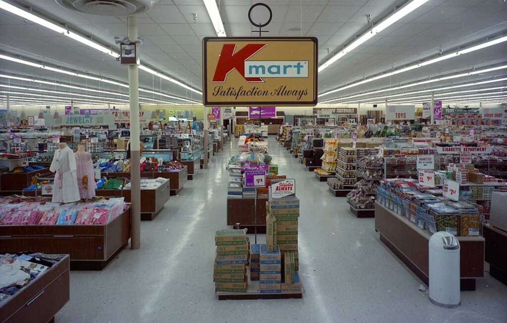 A vintage Kmart store interior with various products displayed, including clothes, electronics, and home goods. The ceiling is lined with fluorescent lights, and the Kmart logo is prominently featured from a suspended sign reading "Satisfaction Always.