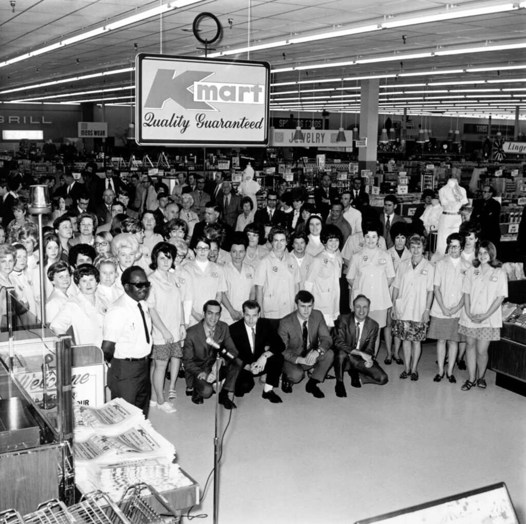 Black and white photo of a large group of Kmart employees posing in a store under a "Quality Guaranteed" sign. The group includes men and women wearing uniforms, with shelves of merchandise visible in the background.