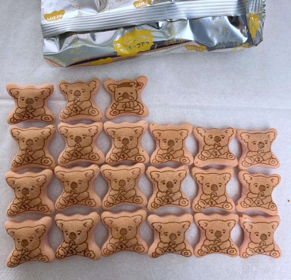 A group of koala-shaped cookies with various printed designs, arranged neatly on a sheet. A partially opened foil package is visible at the top, revealing its contents. The cookies have a light brown color and intricate illustrations.