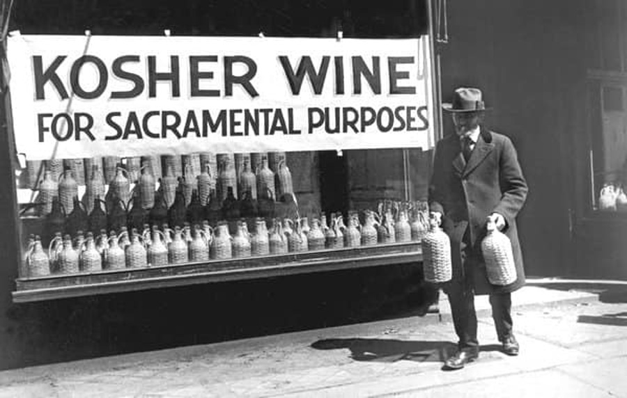 A man in a suit and hat walks on a sidewalk carrying two bottles. Behind him, a shop window displays numerous jugs under a banner reading "Kosher Wine for Sacramental Purposes.