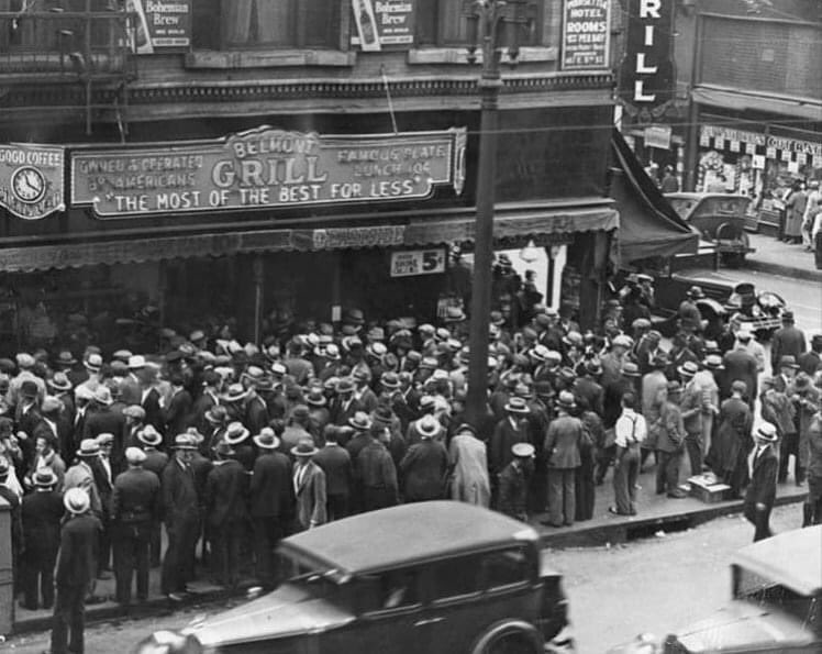 A large crowd of people wearing hats and coats line up outside Belmont Grill under a sign that reads "The Most of the Best for Less." Old-fashioned cars are parked on the street. The scene is black and white, suggesting a historical setting.