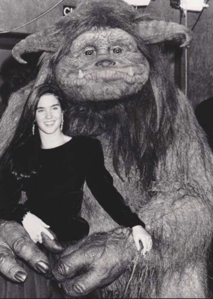 A woman in a black dress smiles while standing next to a large, furry creature with tusks and horns. The creature has long, shaggy hair and rests one hand on the woman's shoulder. The background is indoors with walls and equipment.