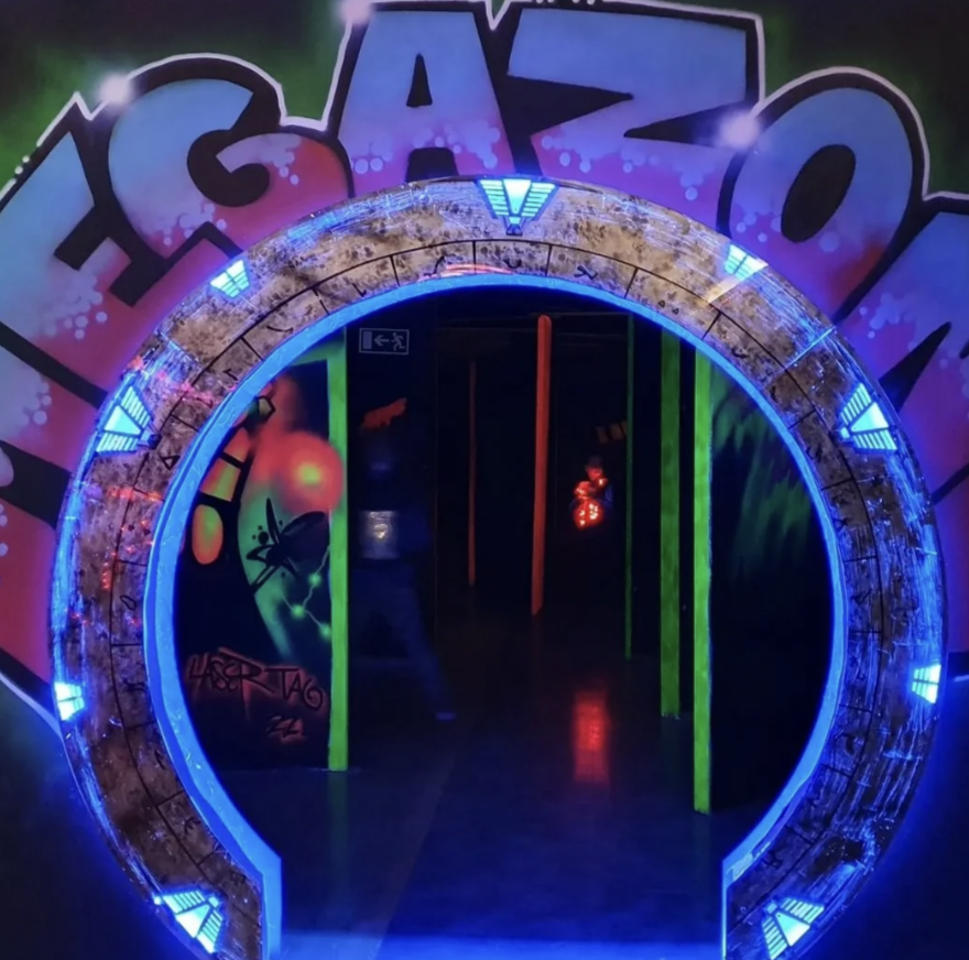 Entrance to a laser tag arena with neon blue lights framing the circular doorway. Colorful graffiti reading "MEGAZONE" is displayed above. The interior is dimly lit with glowing elements.