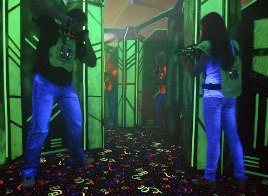 People playing laser tag in a dark, neon-lit arena with glowing walls and a vibrant, patterned floor. Participants are holding laser guns and wearing sensor vests, appearing engaged in the game.